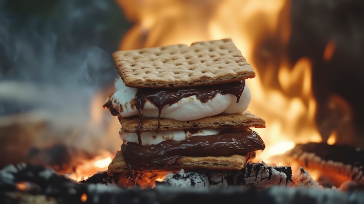 Picture of a pile of graham crackers, melted chocolate and roasted marshmallows in front of a campfire.