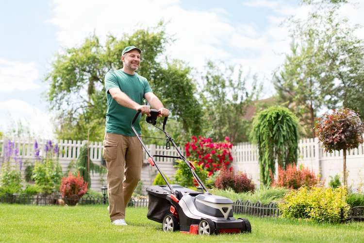 Smiling man is mowing a yard surrounded by flowers and white picket fence.