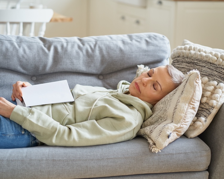 Senior woman napping on a couch with a book on her lap.