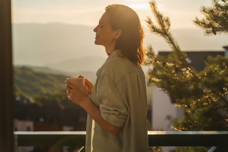 Woman greeting the day with a cup of coffee as she looks out the window at the mountains.