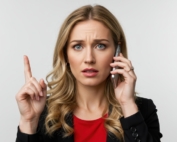 Woman in blazer talking on her cell phone while holding up her pointer finger.