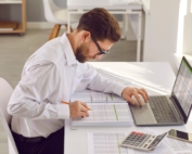 Man in office sitting at a desk, leaning over a long list of detailed pages and his computer.